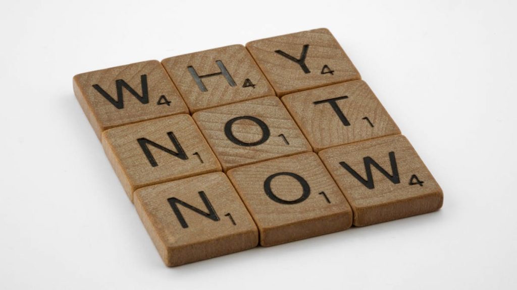Wooden letter tiles form the motivating phrase 'Why Not Now' on a white background, encouraging action and decision-making.